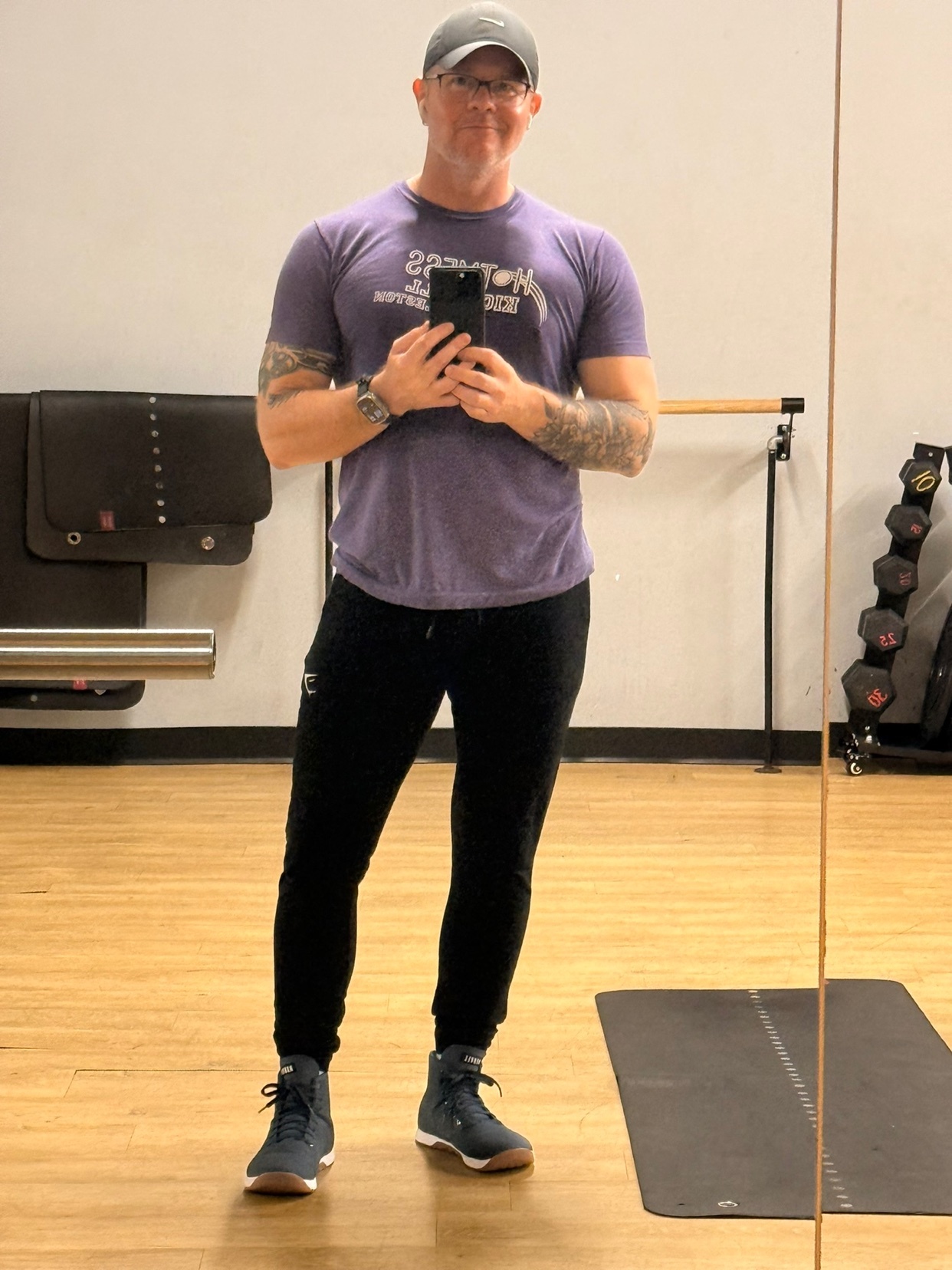 A man in a purple t-shirt and black joggers takes a mirror selfie in a gym. He is wearing glasses and a baseball cap, and has tattoos on his arms.