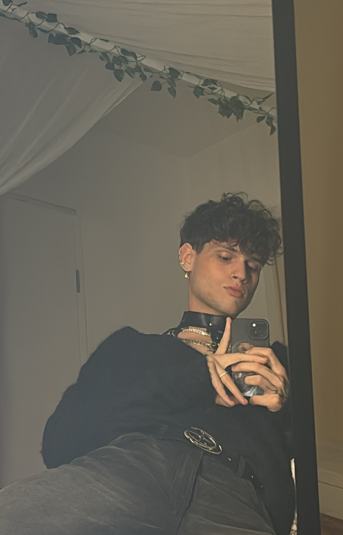 A man takes a mirror selfie, showcasing his style. He is wearing a black top and jewelry.