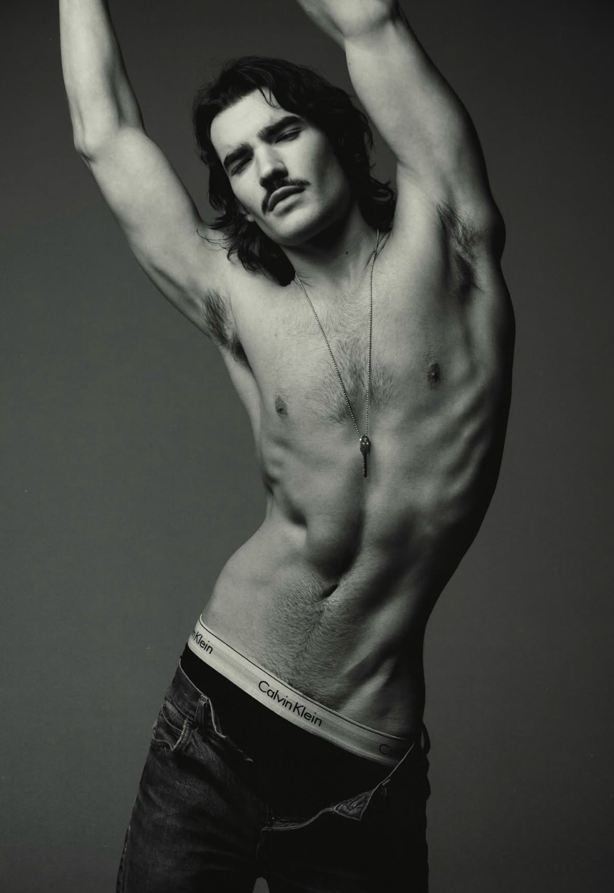 A black and white studio portrait of a shirtless man with a mustache, wearing jeans and Calvin Klein underwear. He has his arms raised above his head and is looking at the camera.