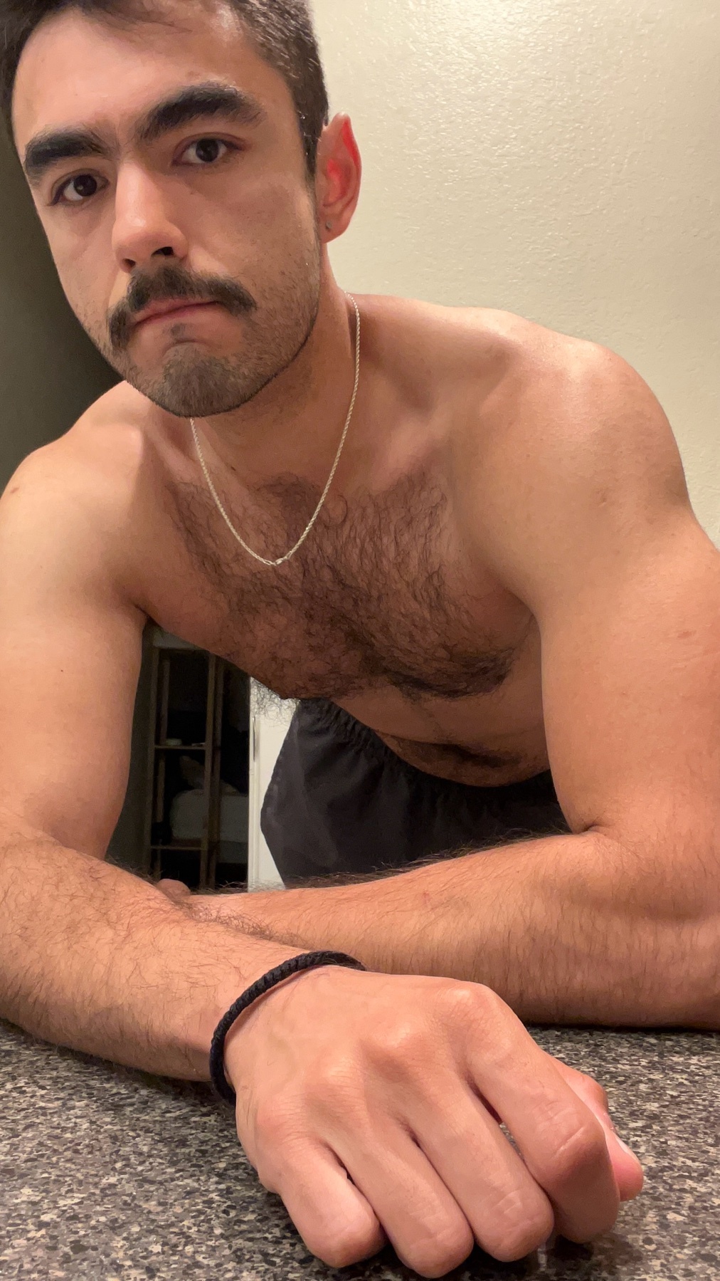 A shirtless man poses for a selfie, showcasing his muscular physique. He has dark hair, a mustache, and a serious expression.