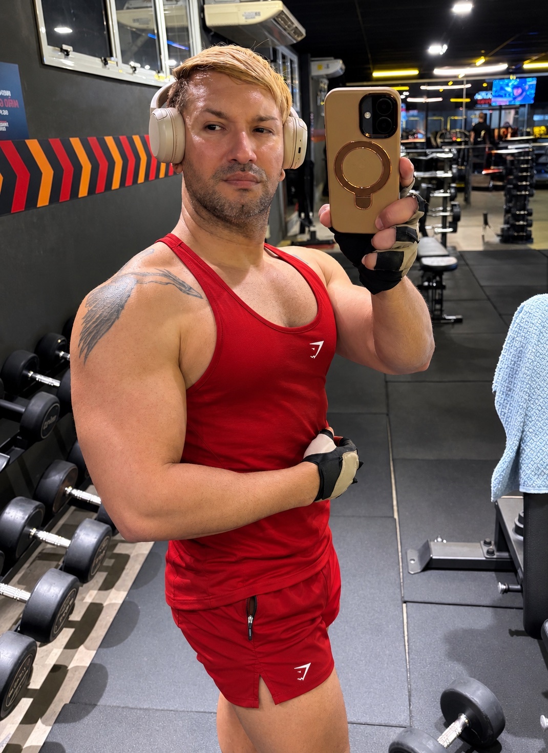 A man with blonde hair and stubble takes a selfie in a gym. He is wearing a red tank top and shorts, and has headphones on.