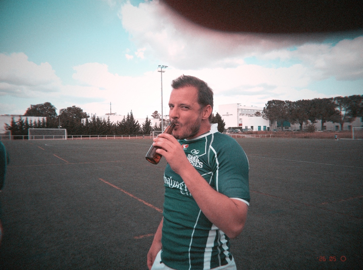 A man in a green rugby jersey drinks a beer on a sports field after a game. The sky is a bright blue with some clouds, and there are trees and buildings in the background.
