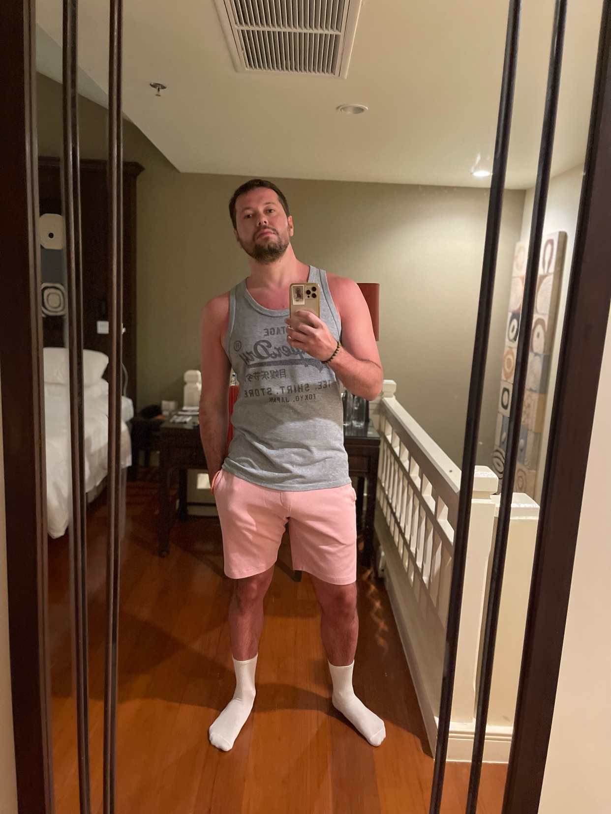 A man takes a mirror selfie, showcasing his outfit. He's wearing a tank top, pink shorts, and white socks.