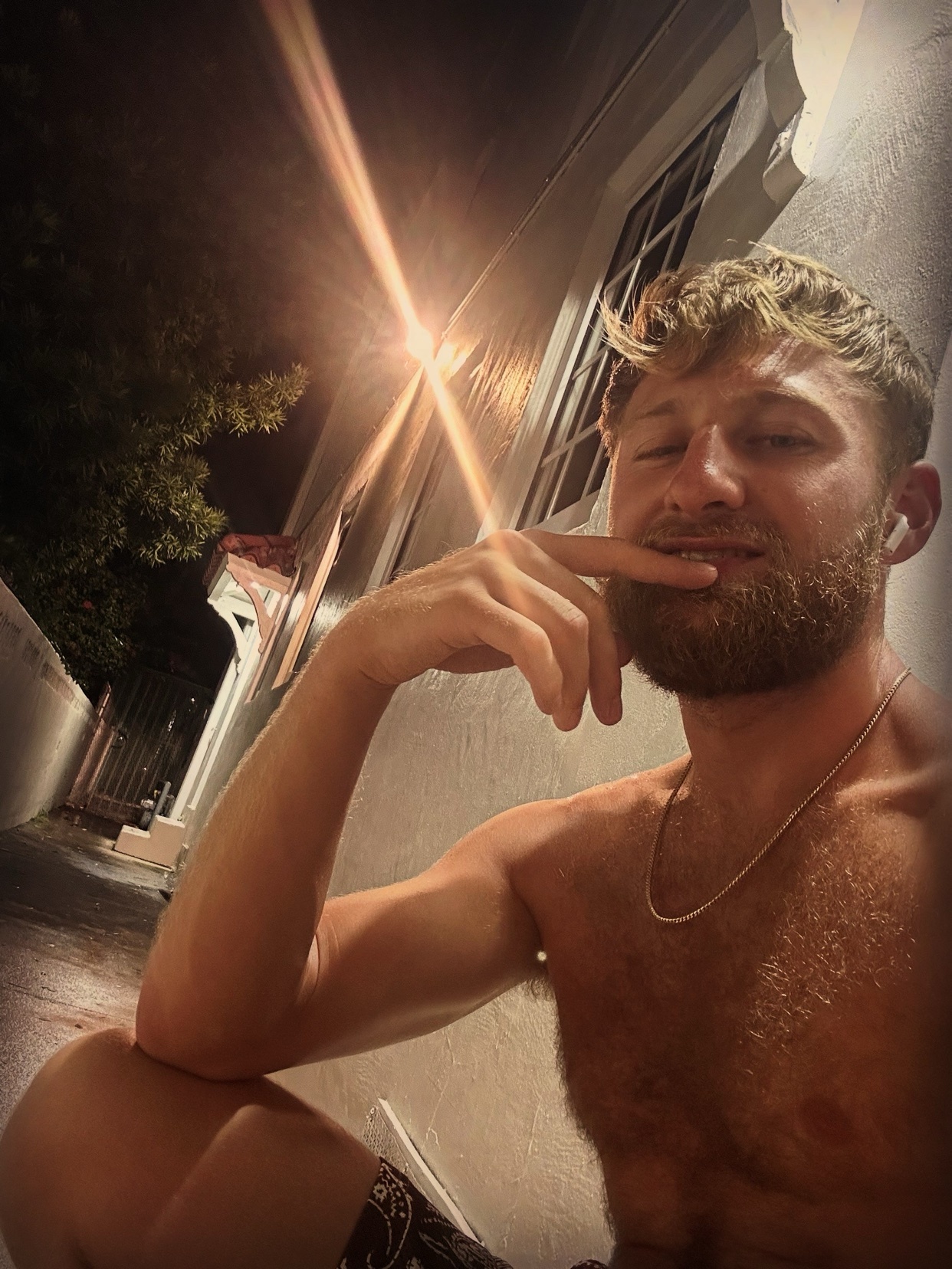 A man with a beard and blond hair poses for a photo at night, wearing earbuds and a necklace. He is shirtless, showing his bare chest and arms.