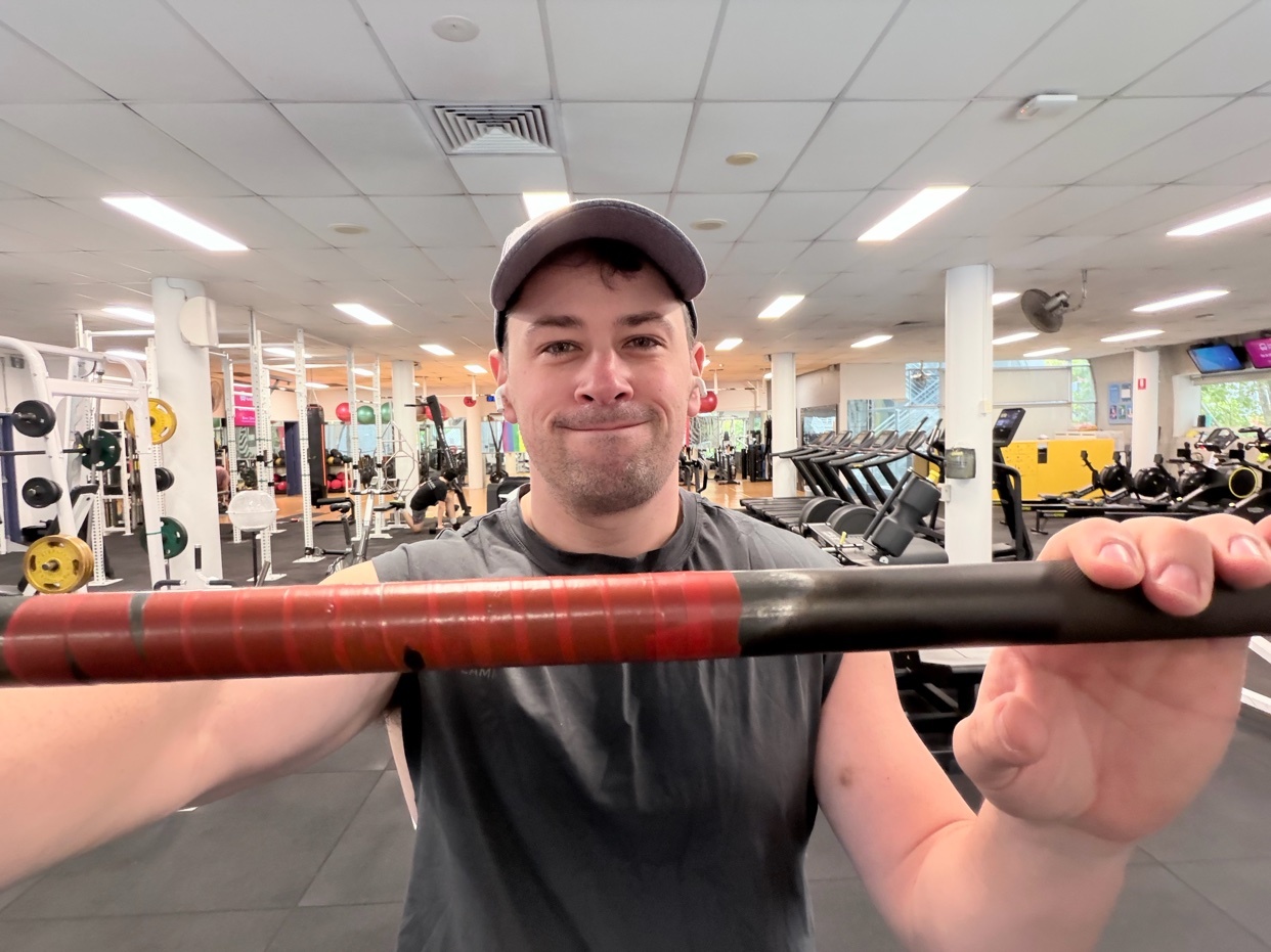 A man in a grey t-shirt and a baseball cap smiles at the camera while holding a barbell in a gym. The barbell has red tape wrapped around the middle section.