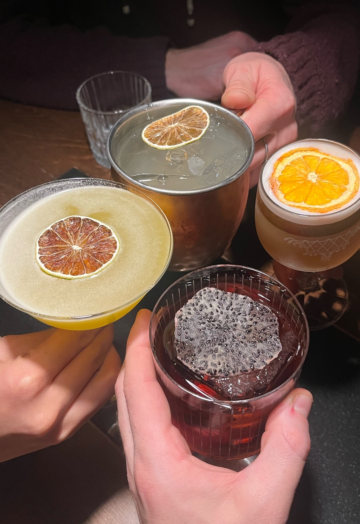 A close-up shot of a variety of cocktails, each garnished with a slice of fruit. The drinks are held by hands, suggesting a social gathering.