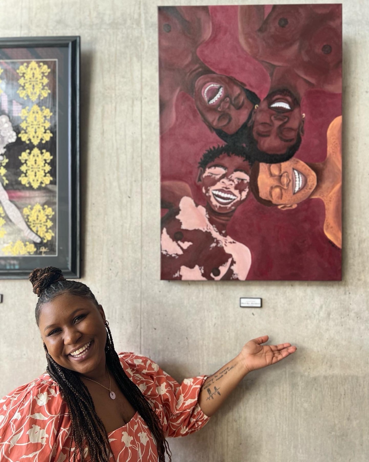 A woman with braided hair and a floral top smiles and gestures towards a painting on a wall. The painting depicts a close-up, upside-down view of four laughing Black men.