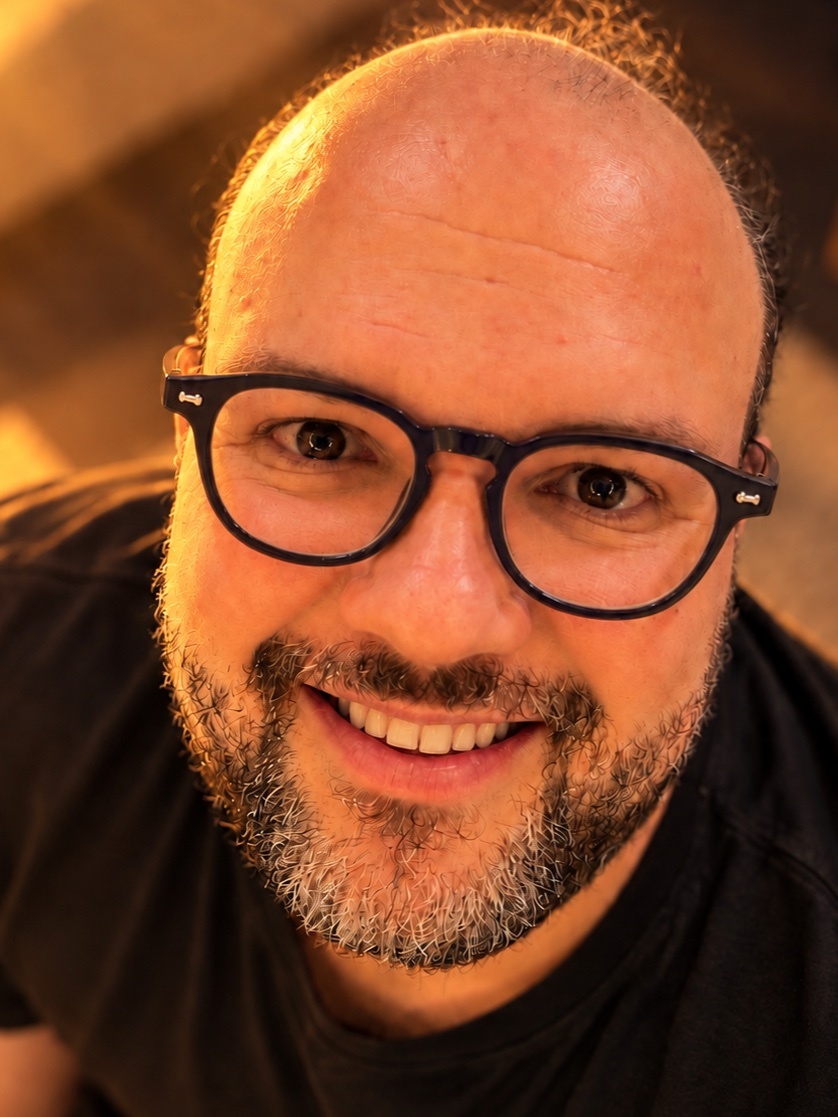 A close-up shot of a smiling man wearing black-rimmed glasses and a beard. The man is bald with some hair around the sides of his head.