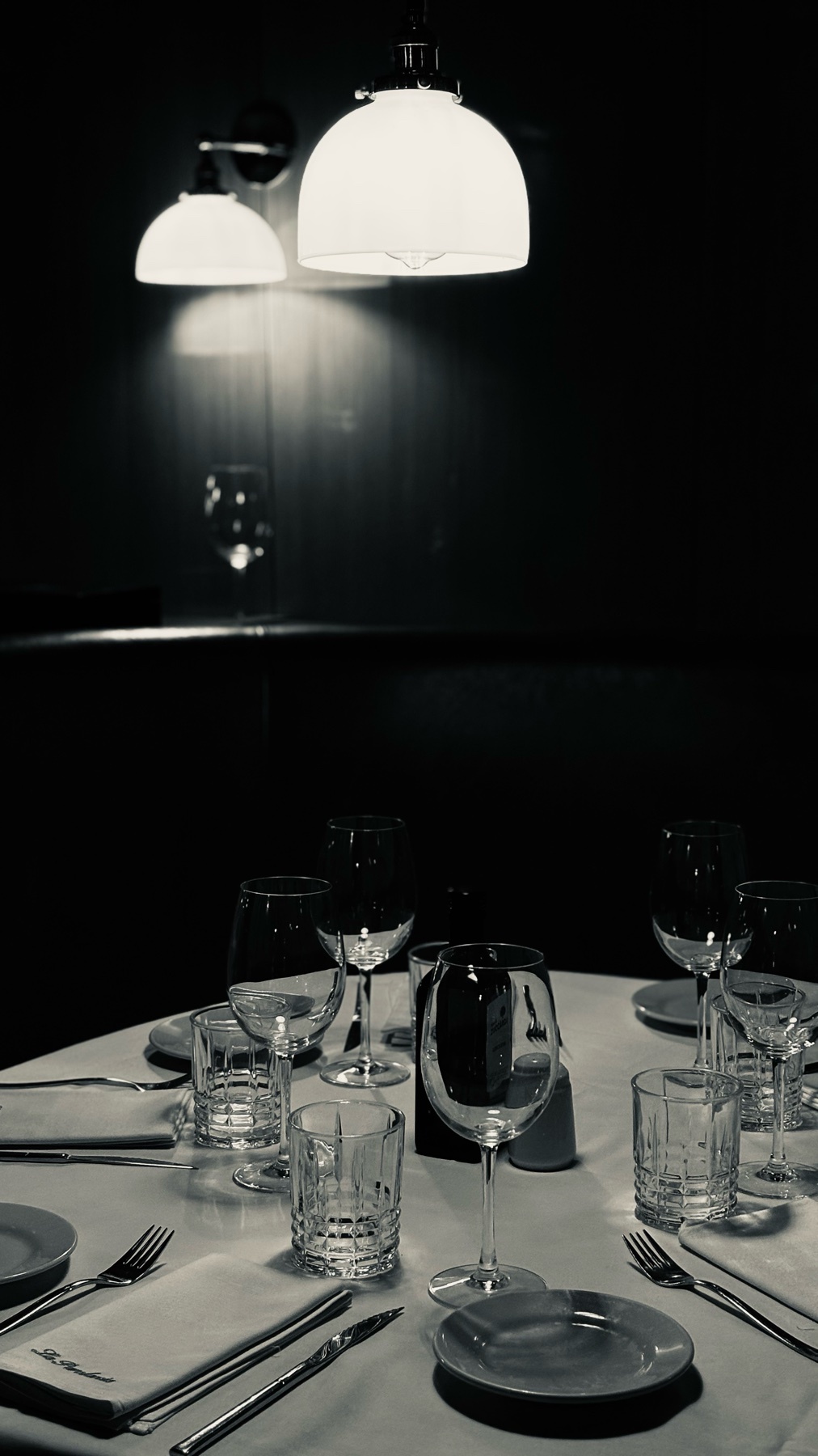 A black and white photo of a restaurant table setting. The table is set with glasses, silverware, and plates under soft lighting.
