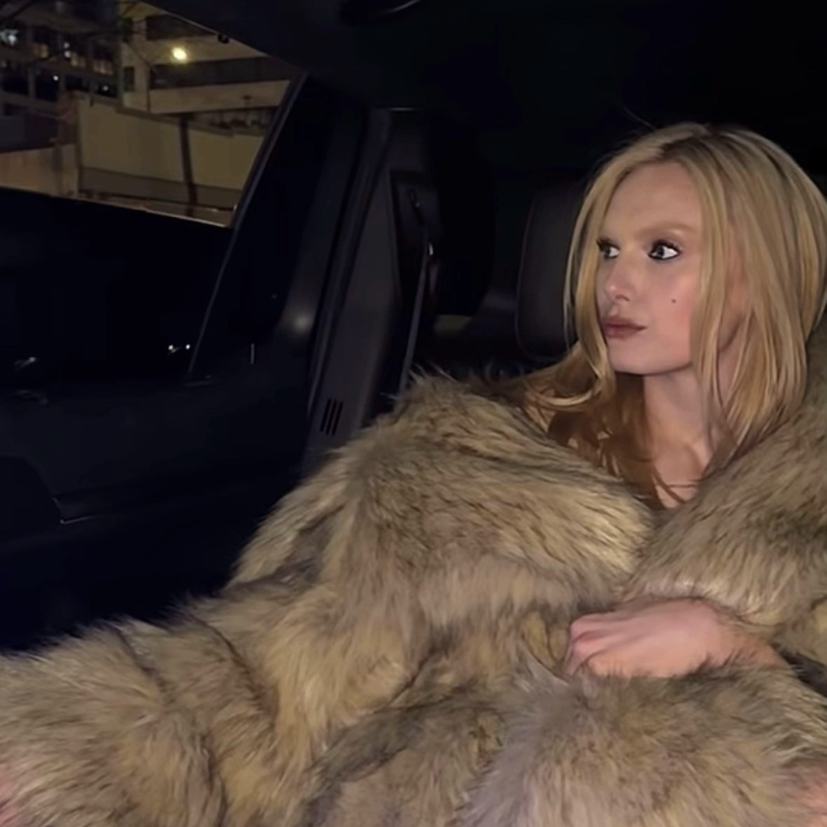 A woman with blonde hair wearing a fur coat sits in a car at night. The photo has a glamorous and stylish aesthetic.