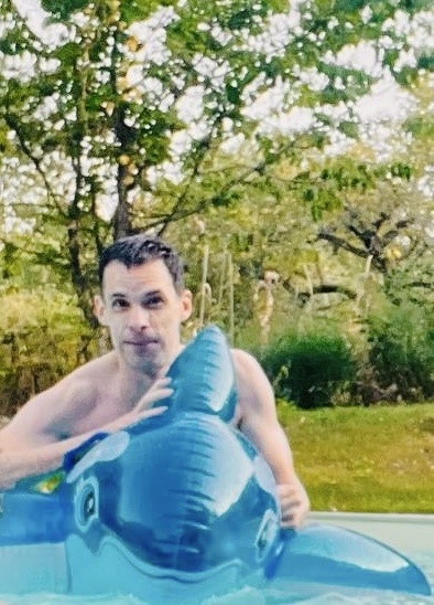 A man is in a pool, holding an inflatable whale. He is shirtless and looking at the camera.