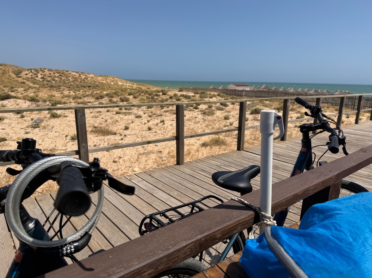 Two bicycles are parked on a wooden boardwalk overlooking sand dunes and the ocean on a sunny day.