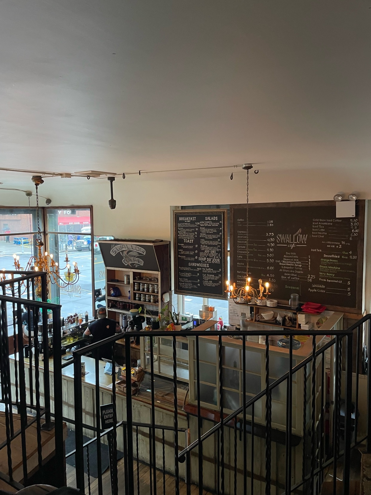 A view from above shows the interior of a cozy coffee shop with two large menu boards on the wall. A barista is visible behind the counter, and decorative chandeliers hang from the ceiling.