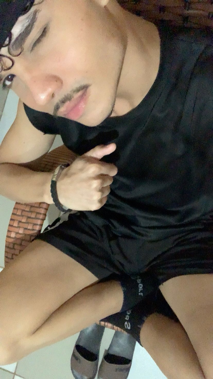 A man with curly hair and a mustache poses for a selfie, wearing a black t-shirt and shorts.