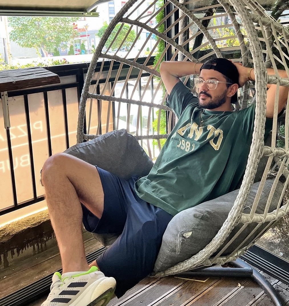 A man with a beard and glasses sits in a hanging chair, wearing a green t-shirt and navy shorts. He has his hands behind his head and appears relaxed.