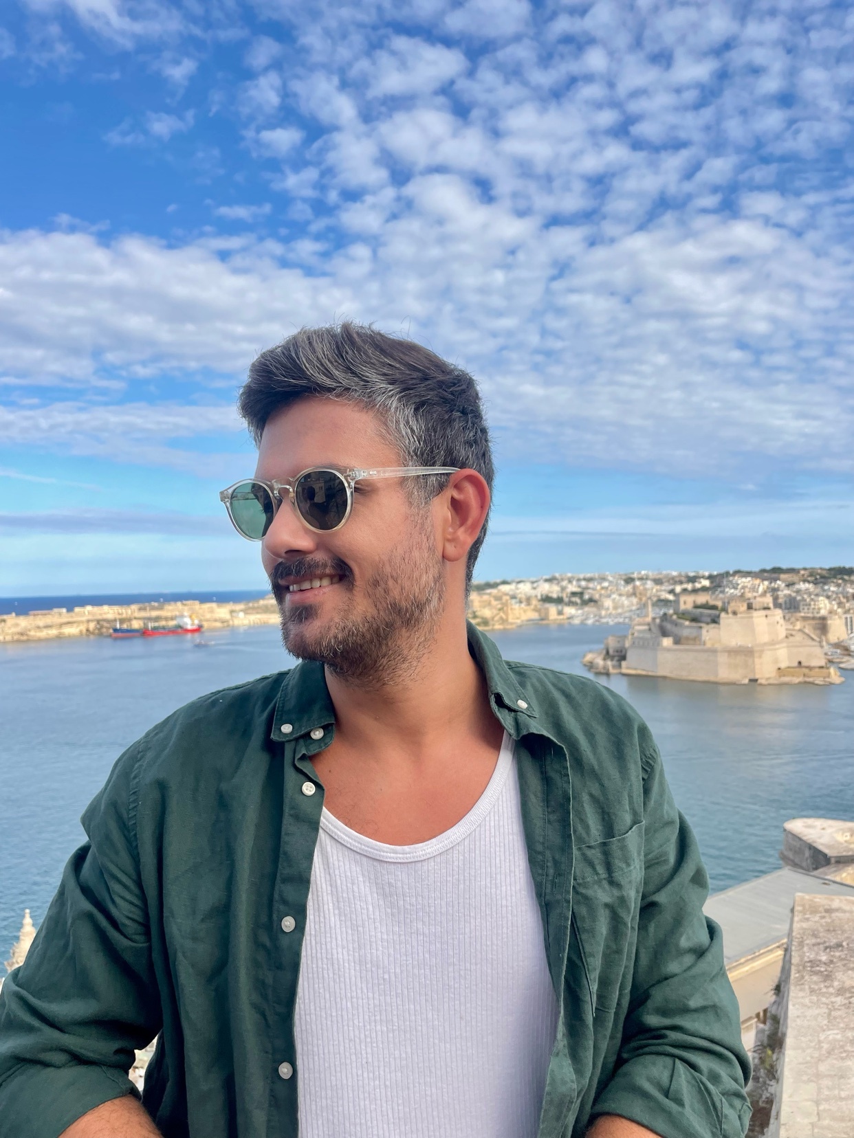 A man with sunglasses and a beard smiles as he looks to the side with a European cityscape and blue sky in the background.