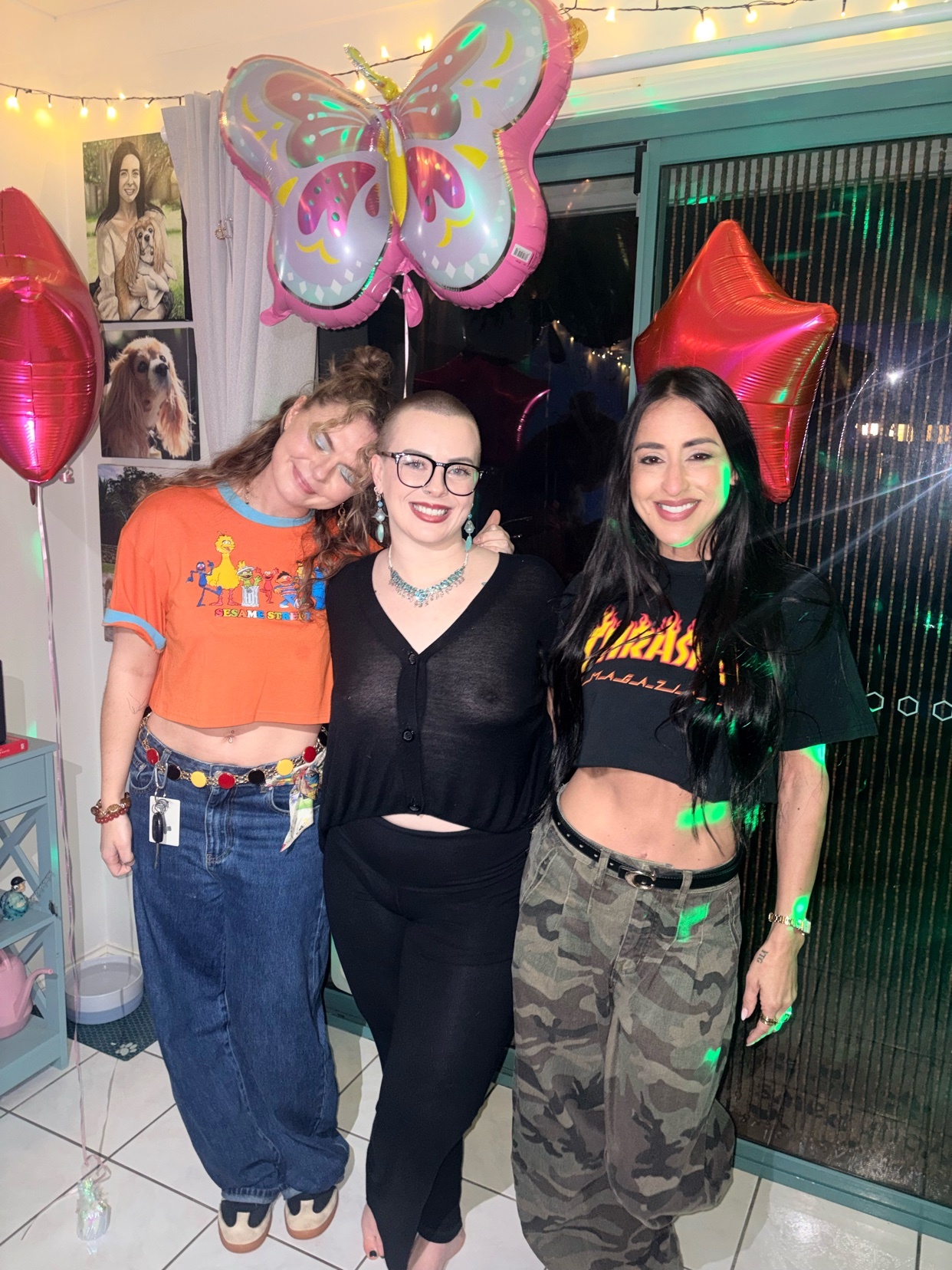 Three women pose for a photo at a party, surrounded by balloons and string lights. The woman on the left wears an orange graphic t-shirt and baggy jeans, the woman in the middle wears a black cardigan and leggings, and the woman on the right wears a