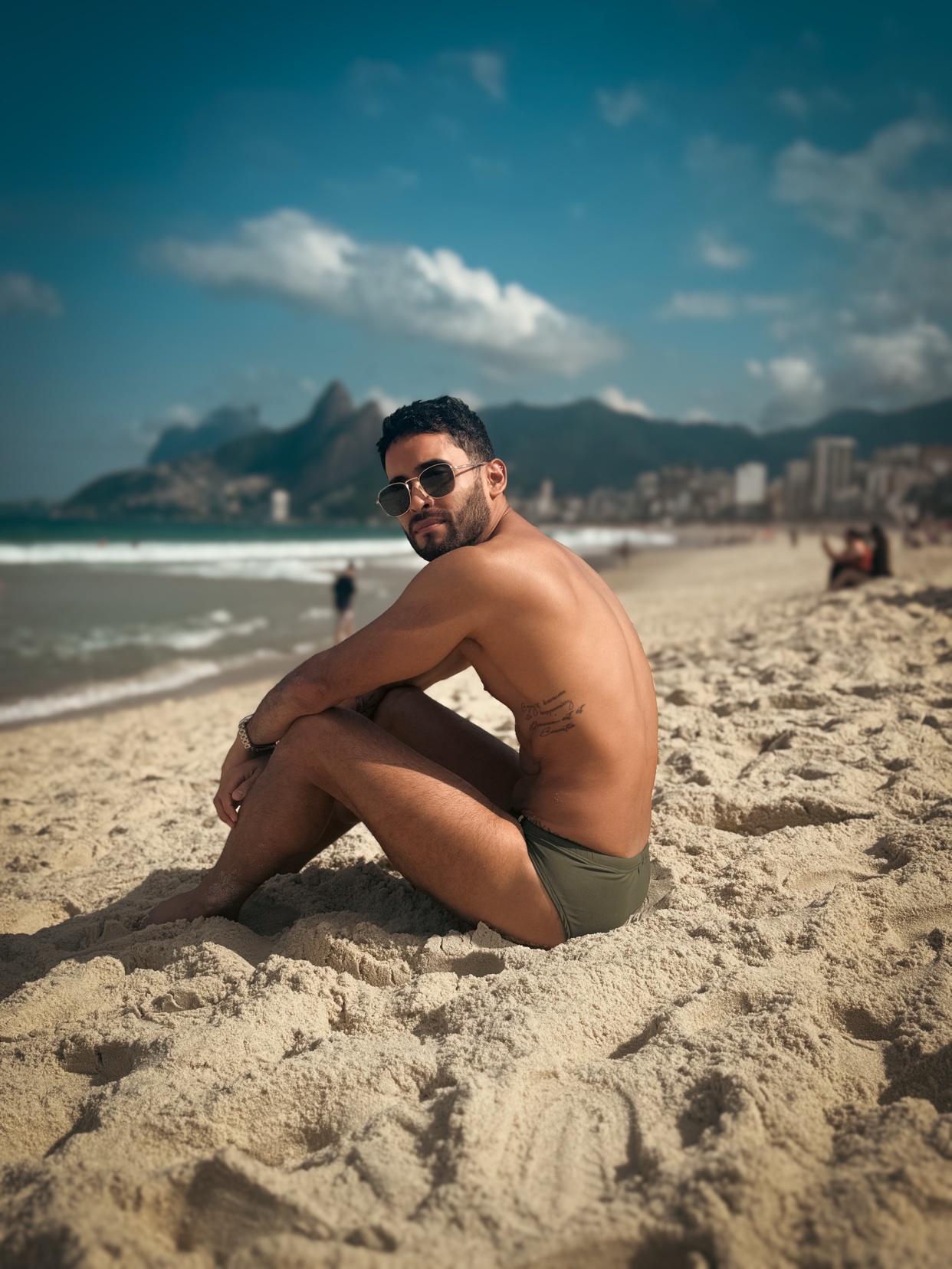 A man with sunglasses and a tattoo sits on a sandy beach, looking over his shoulder towards the ocean. Mountains and a cityscape are visible in the background under a bright blue sky.