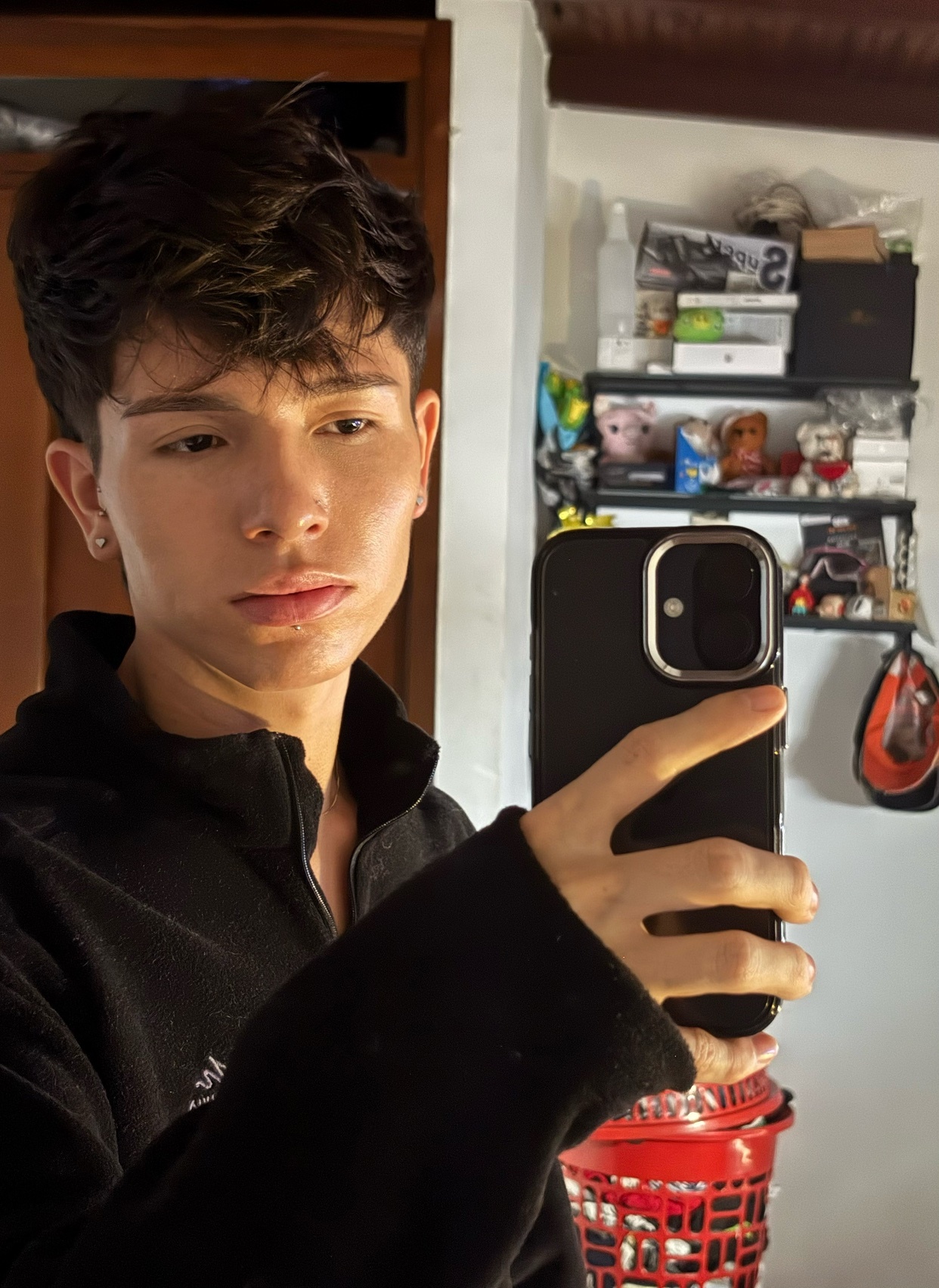 A young man with dark, curly hair takes a selfie in a black fleece jacket. He is looking towards the camera with a neutral expression.