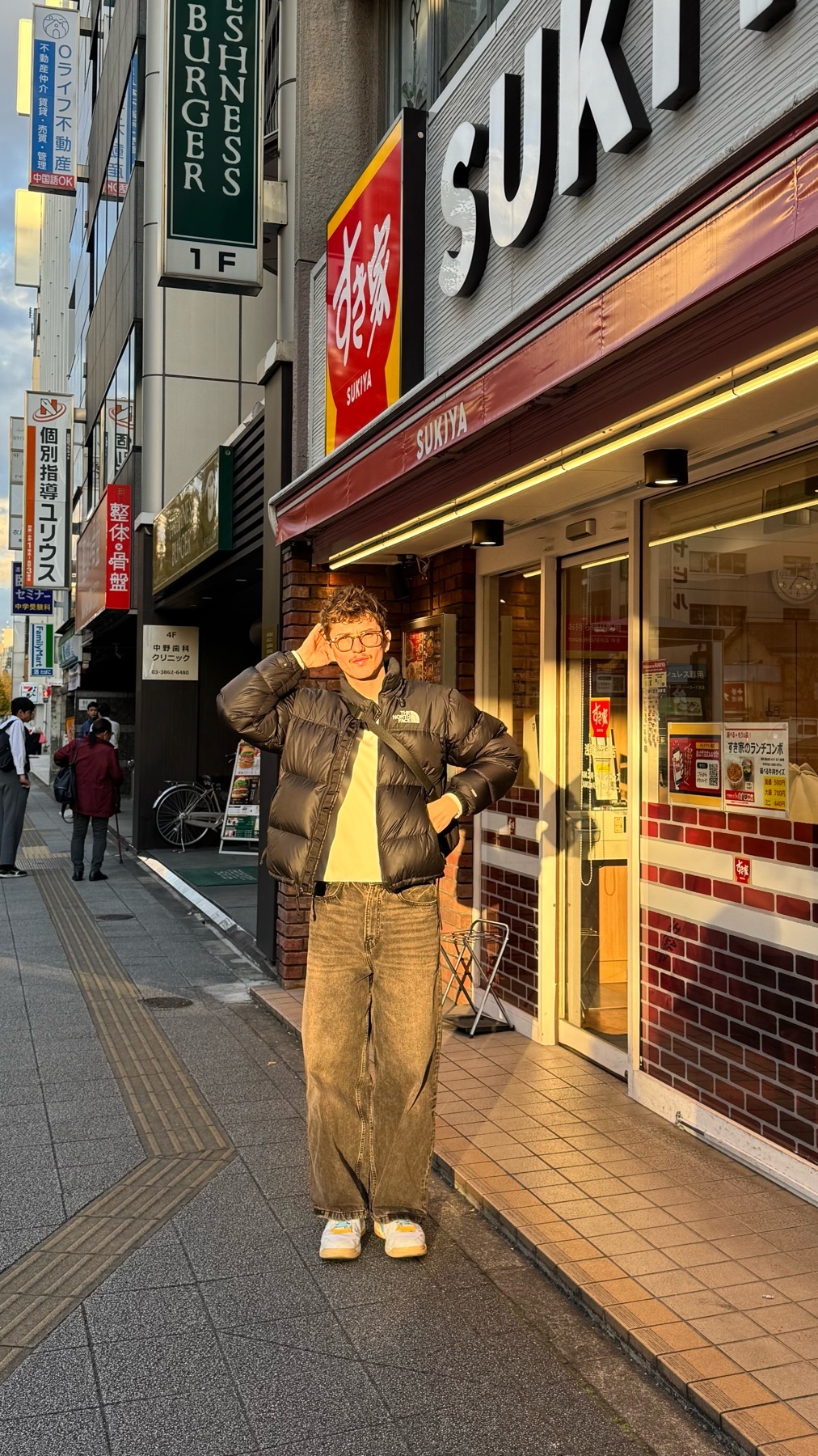 A man poses in front of a Japanese restaurant. He is wearing a puffer jacket, jeans, and sneakers.