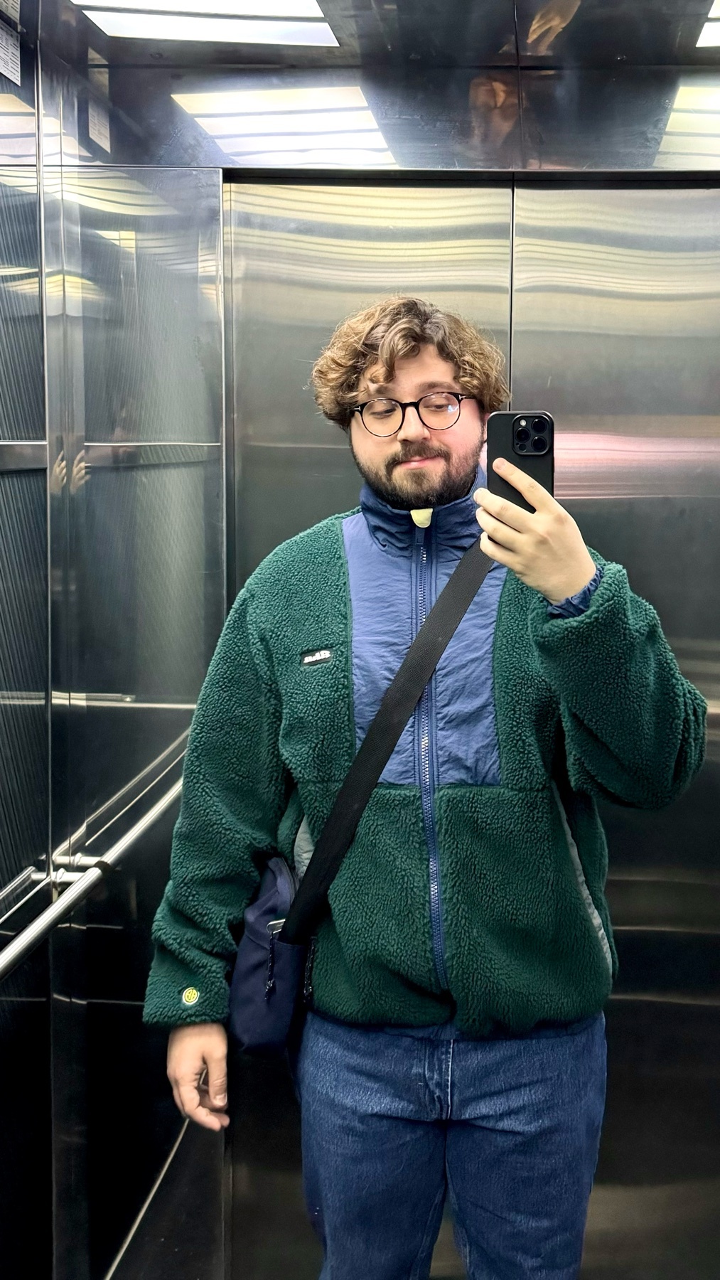 A man takes a selfie in an elevator. He is wearing a green and blue jacket, glasses, and jeans.