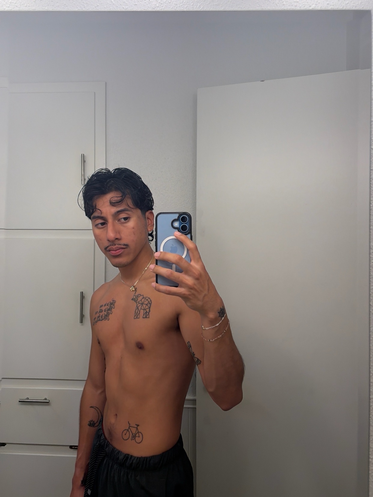 A man takes a mirror selfie, shirtless, showing off his tattoos and physique. He has dark hair and a mustache.