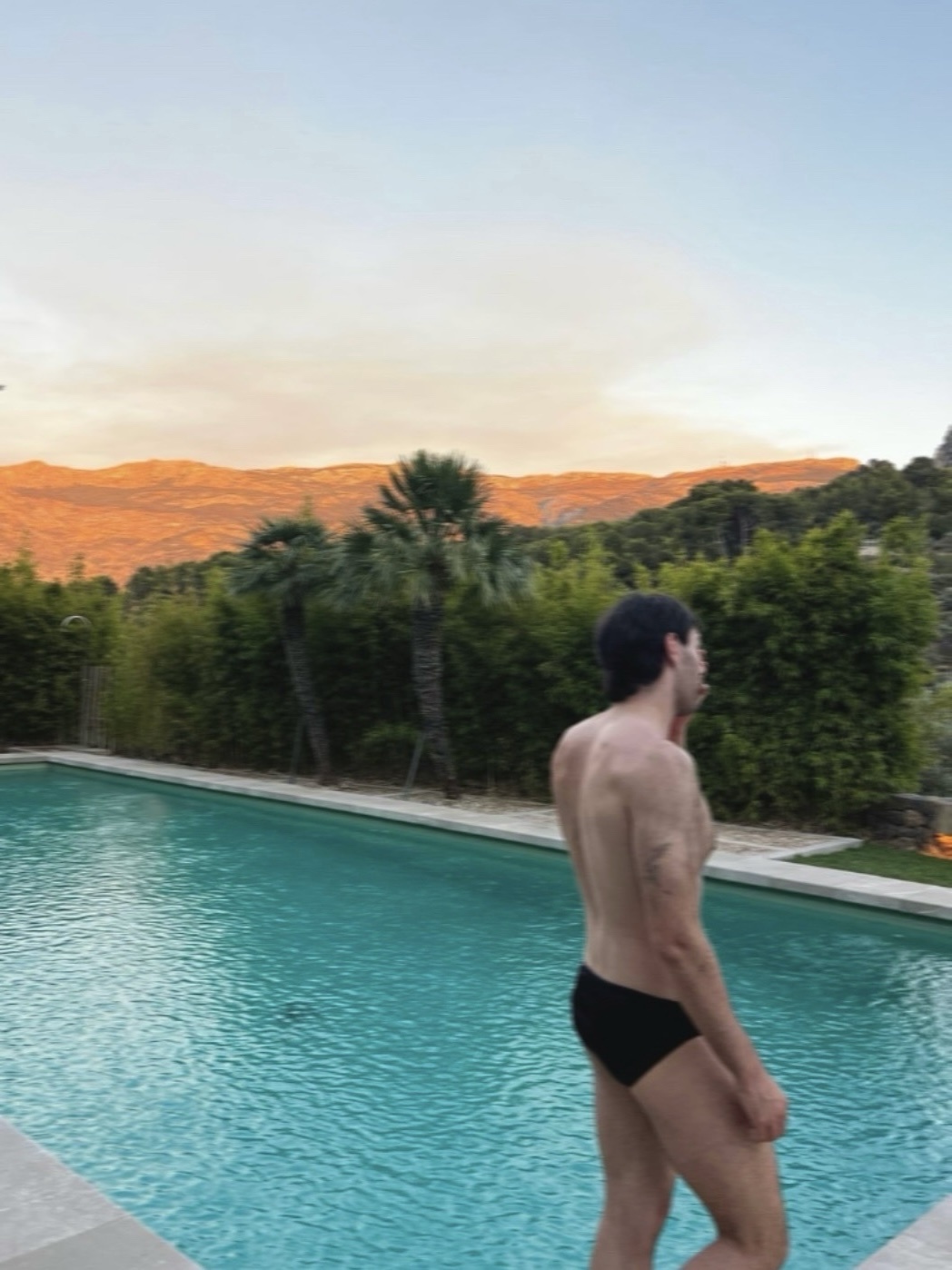 A man in black swimming trunks stands by a pool with mountains in the background at sunset.