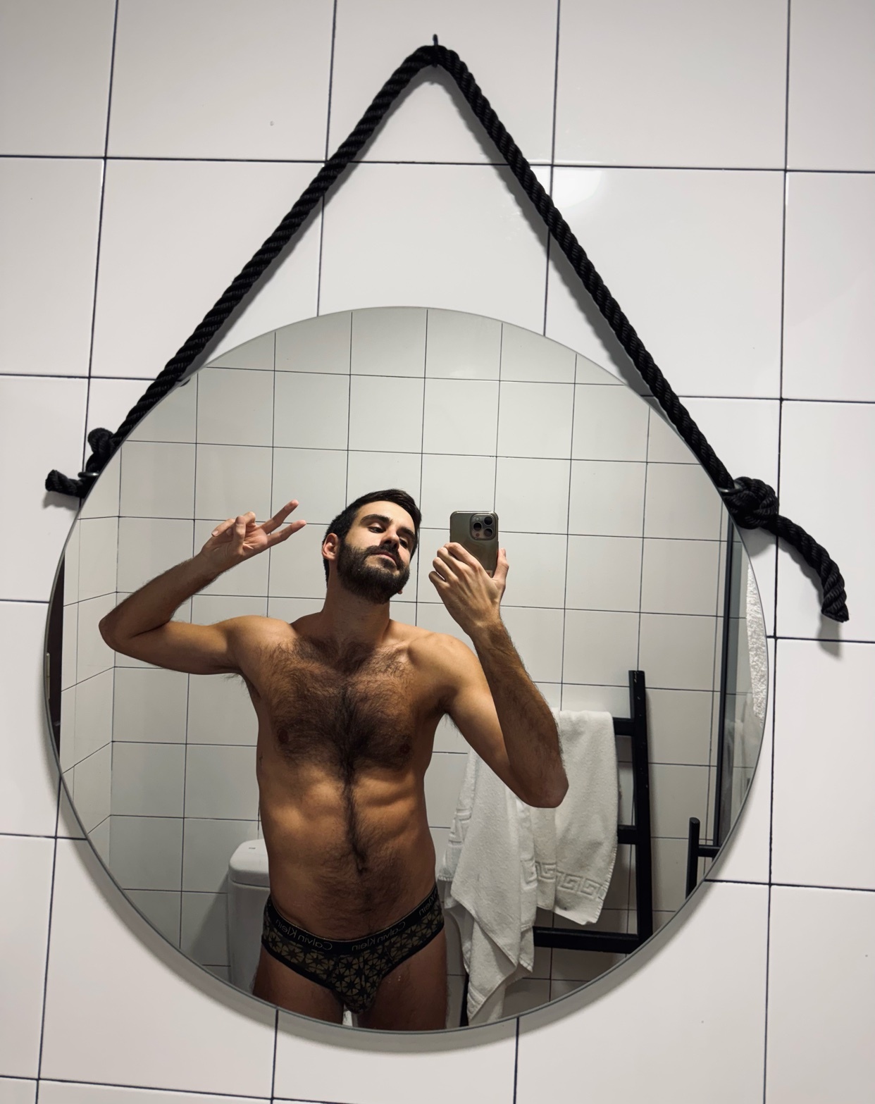 A man takes a mirror selfie, shirtless and wearing patterned underwear. He's in a bathroom with a round mirror and a black rope hanging it.