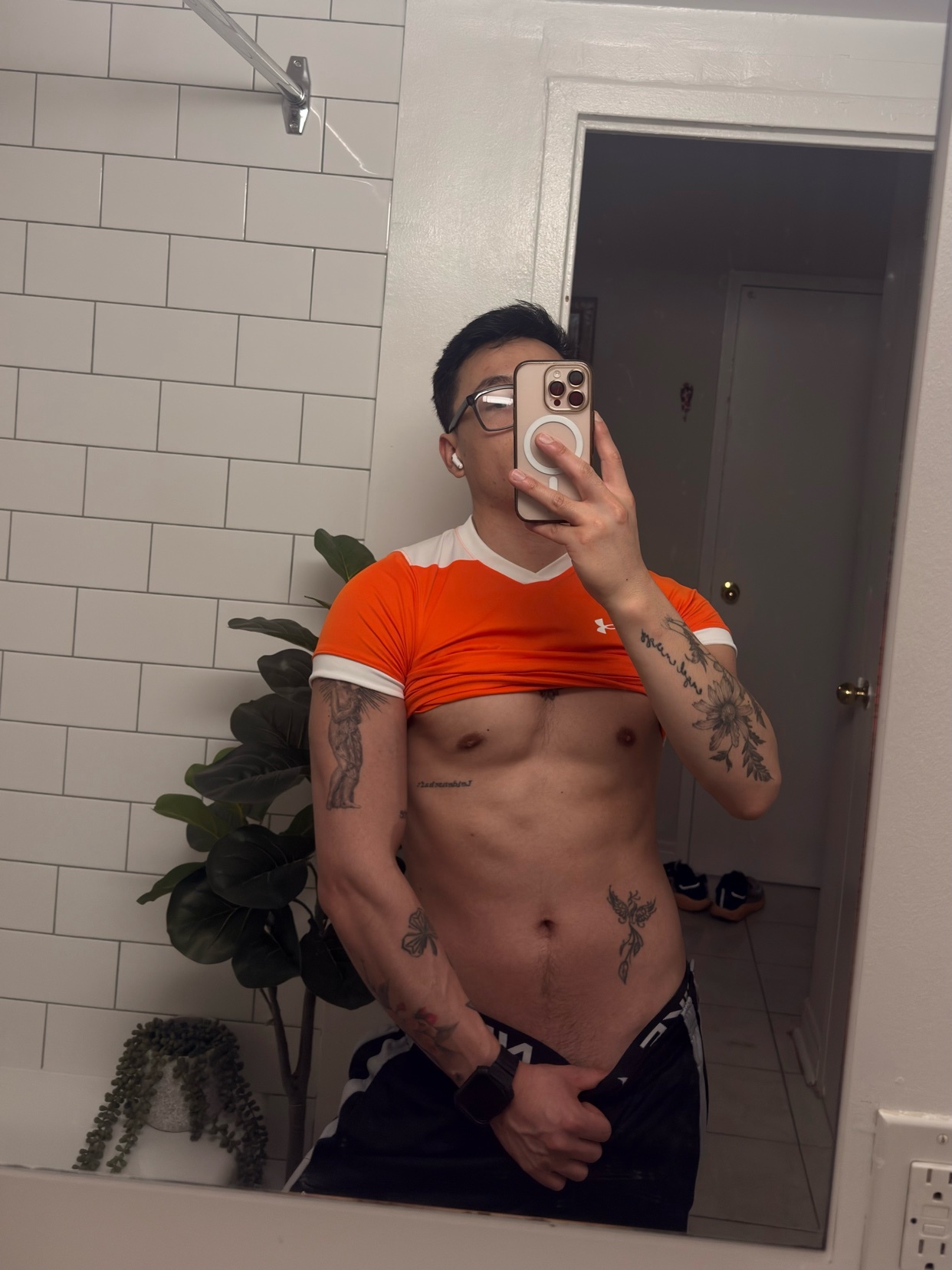 A man with tattoos and glasses takes a mirror selfie in a bathroom. He is wearing an orange crop top and black shorts, and his torso is bare.
