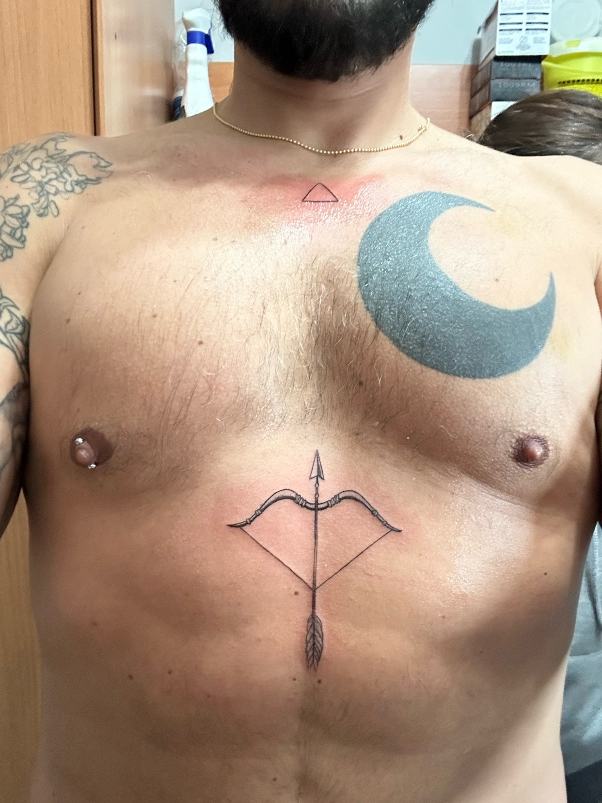A man shows off his chest tattoos, including a bow and arrow, a crescent moon, and a small triangle. He also has nipple piercings.