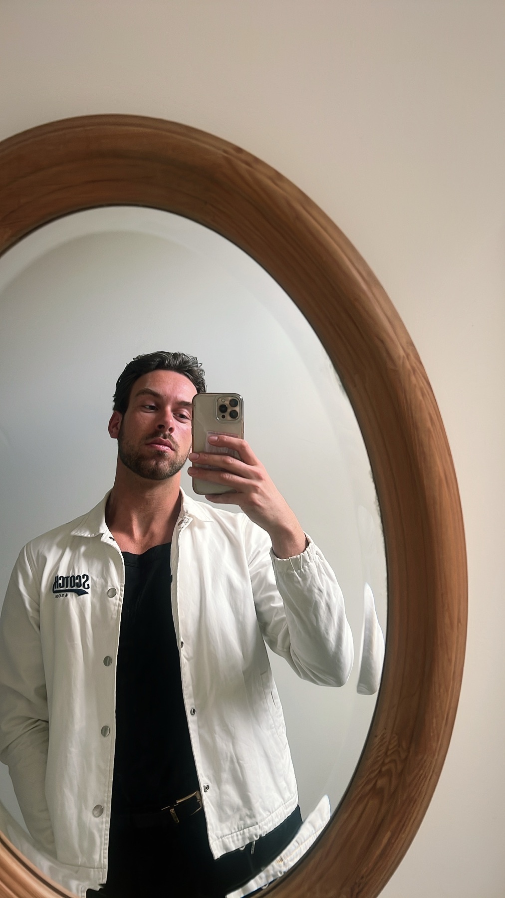 A man takes a mirror selfie while wearing a white jacket over a black t-shirt. He has dark hair and stubble.