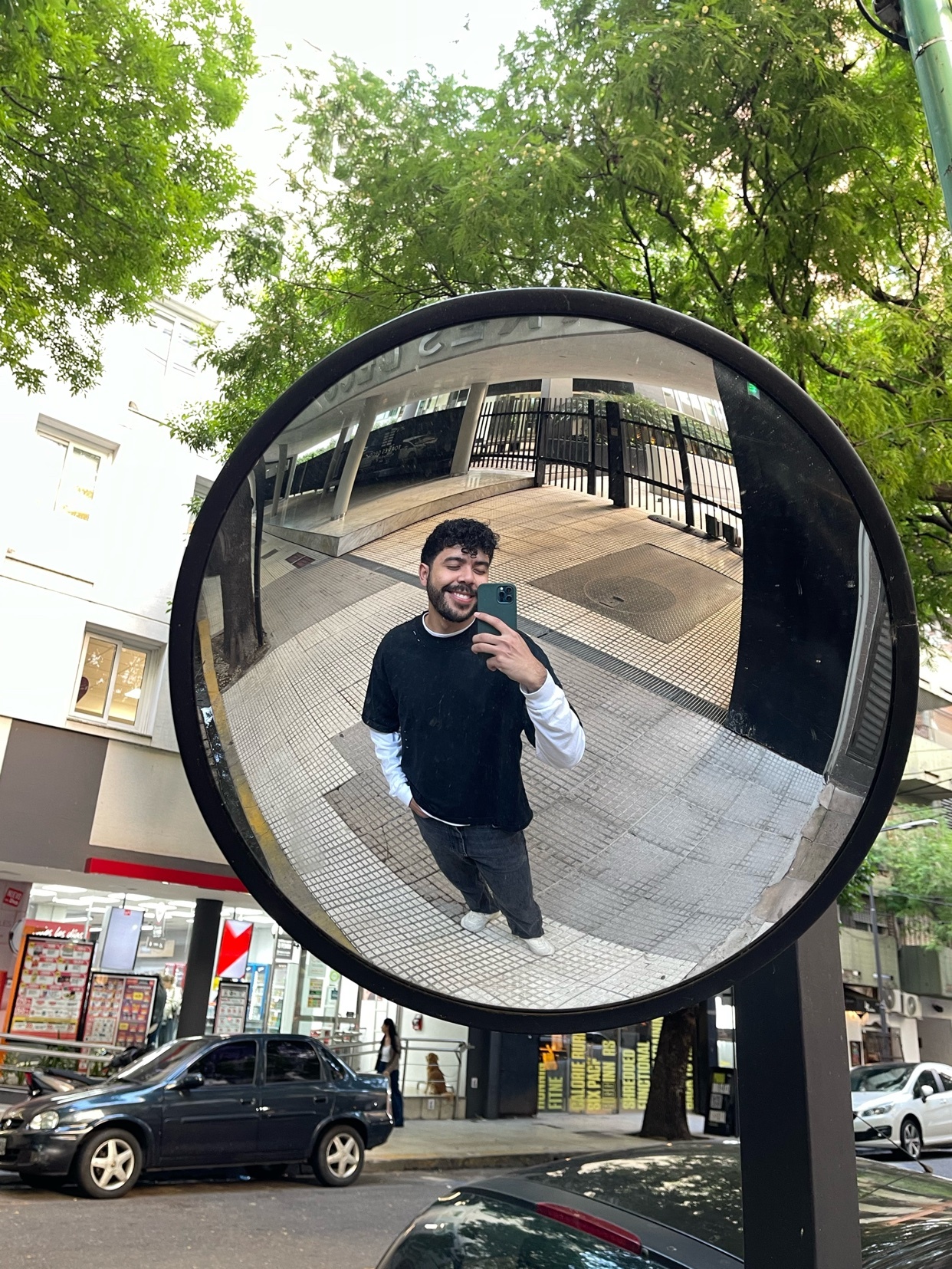 A man takes a selfie in a large, round mirror on a city street. The reflection captures the urban environment and the man's smiling face.