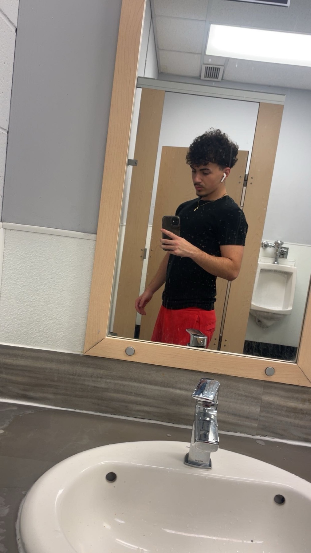 A man takes a mirror selfie in a public restroom. He is wearing a black t-shirt and red shorts, with earbuds in his ears.