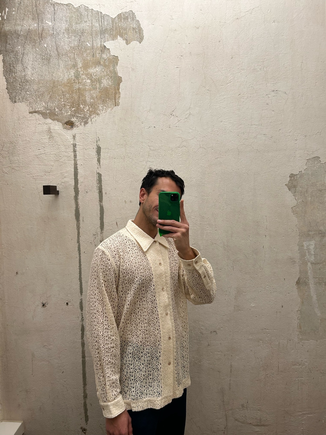 A man takes a selfie in a cream-colored, long-sleeved lace button-up shirt. He is holding a green phone in front of his face, obscuring it. The background is a textured, off-white wall with peeling paint.