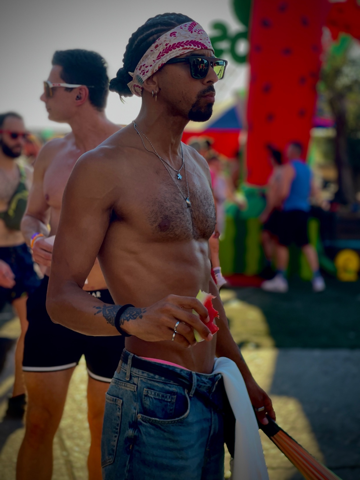 A shirtless man with braids and a bandana tied around his head, wearing sunglasses and denim shorts, holds a slice of watermelon. He is at an outdoor event with other people in the background.