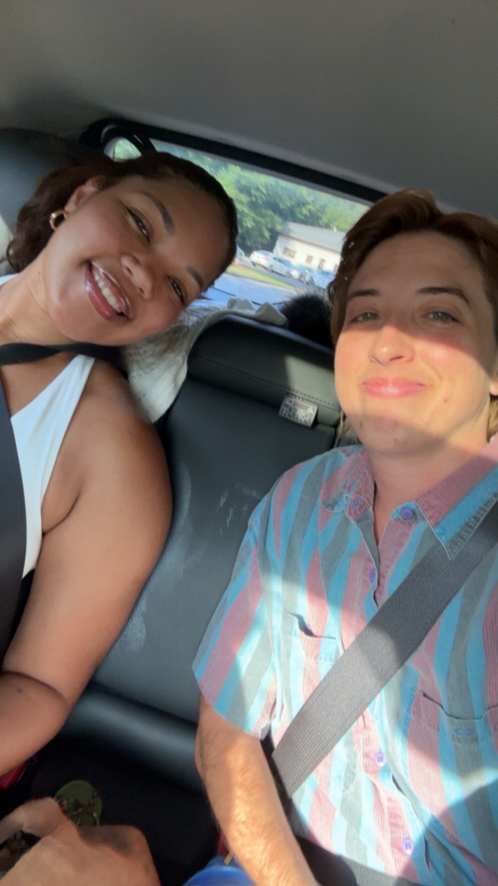A smiling couple is captured in a selfie taken from the back seat of a car. The woman on the left is wearing a white tank top, and the person on the right is wearing a colorful striped button-down shirt.