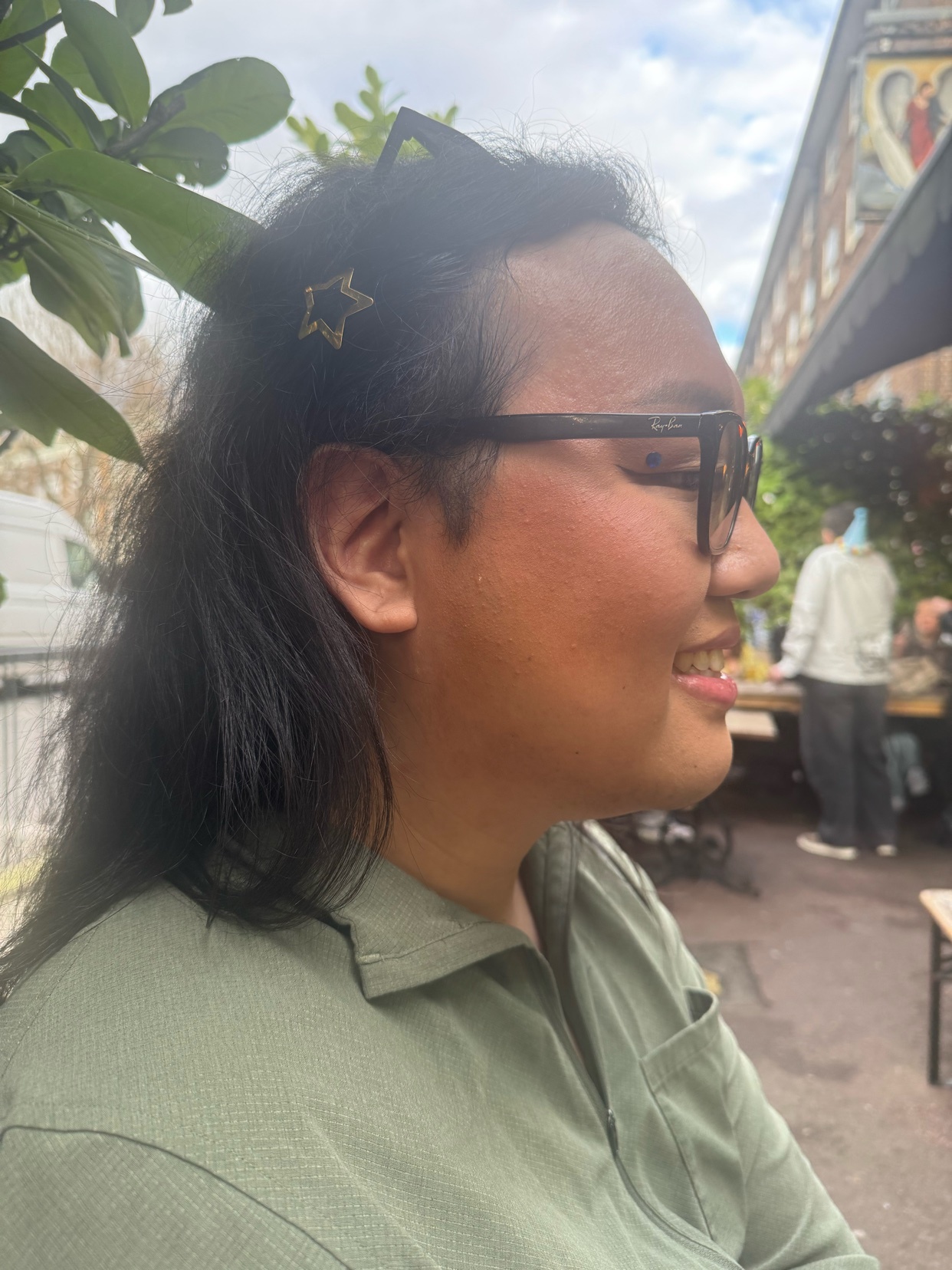 A woman with dark hair and glasses smiles while sitting outdoors. A star-shaped hair clip is visible in her hair.