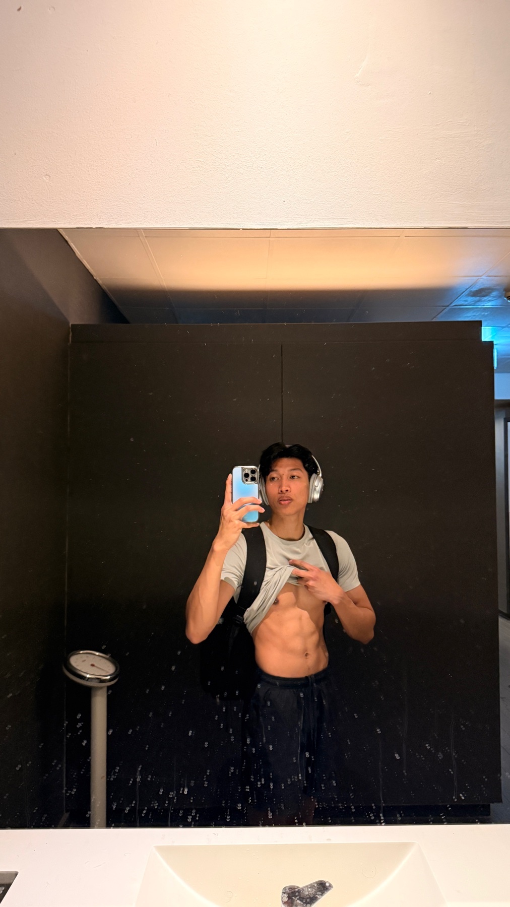 A young Asian man with defined abs lifts his shirt to show his midsection while taking a mirror selfie in a gym. He is wearing headphones and a backpack.