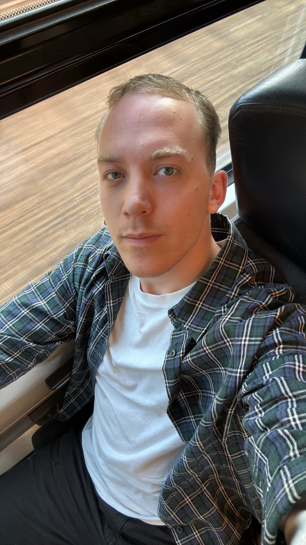 A man takes a selfie while sitting down. He is wearing a plaid shirt over a white t-shirt.
