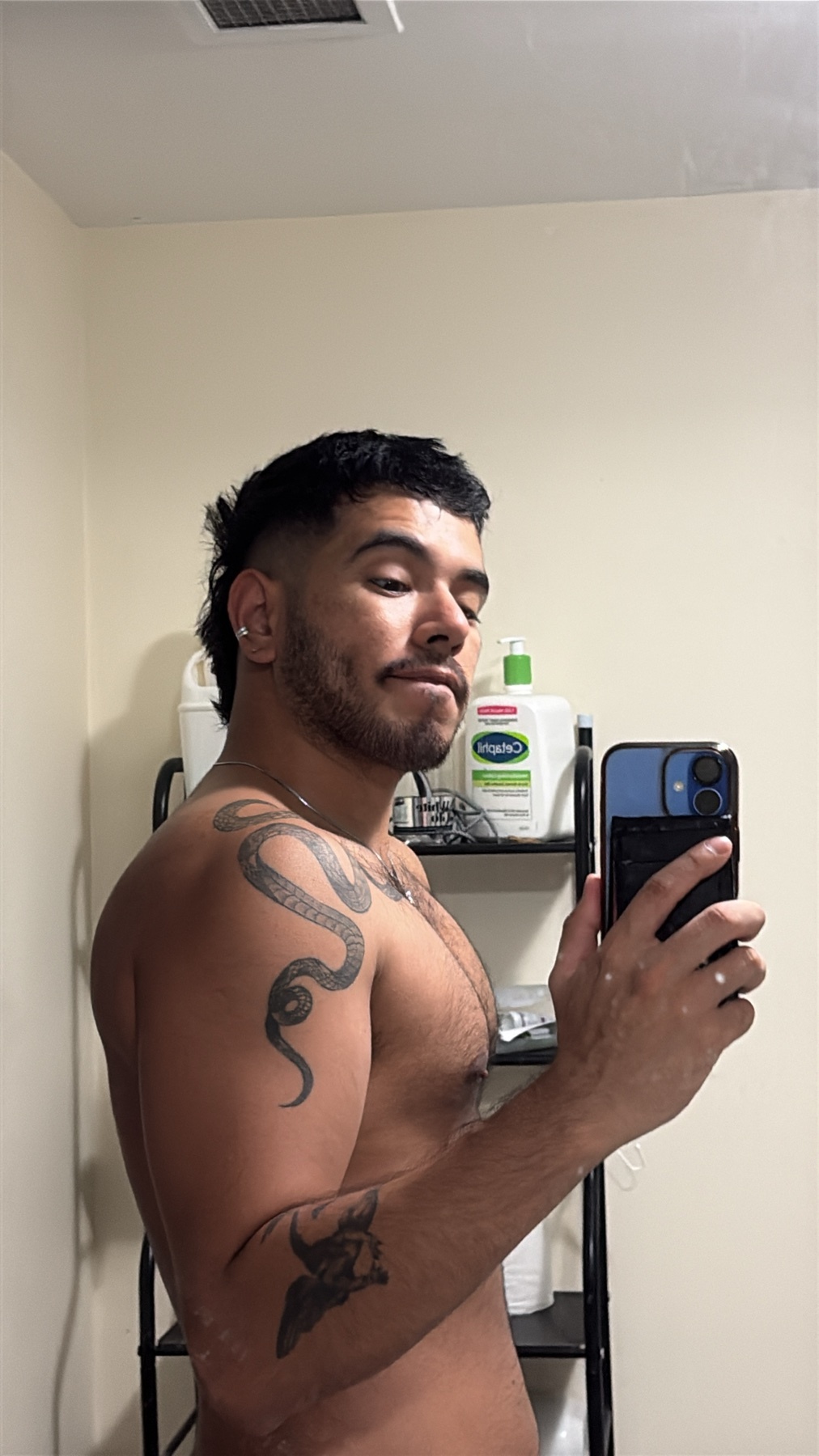 A man takes a mirror selfie, shirtless, showing off his tattoos and physique. The photo is taken in a bathroom.