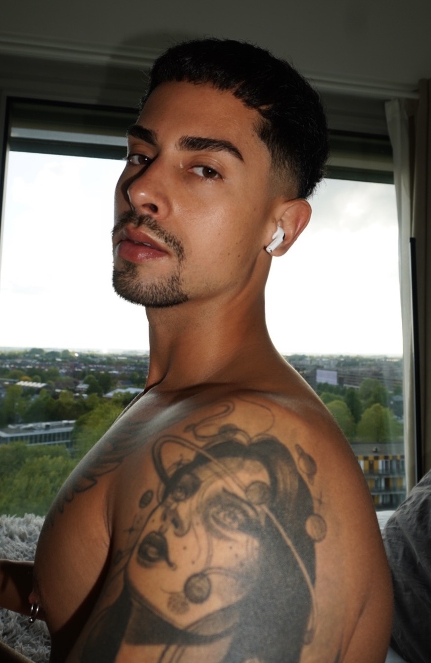 A man with dark hair and stubble poses for a portrait, showing off a large tattoo on his shoulder and chest. He is wearing an earring and has a piercing on his nipple.