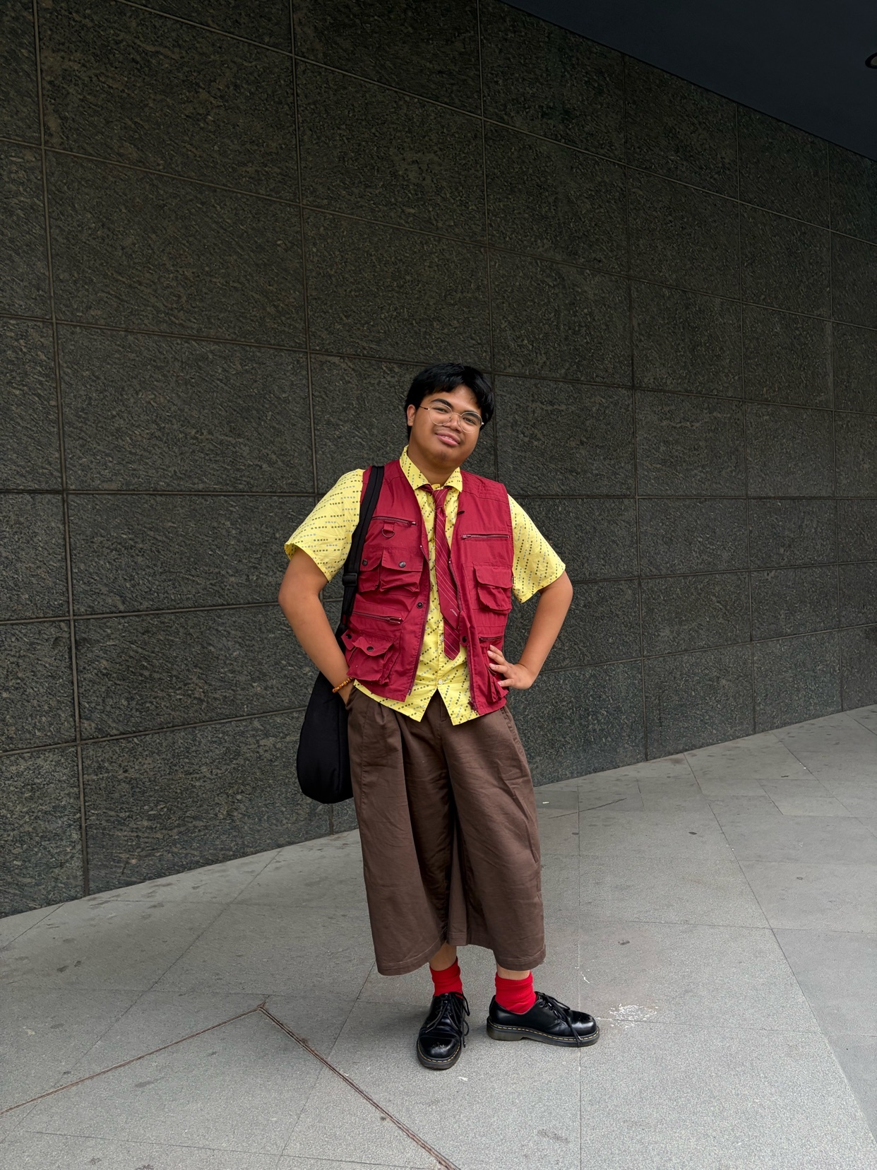 A man poses in a fashionable streetwear outfit featuring a yellow button-up shirt, a red vest, brown wide-leg pants, and red socks.