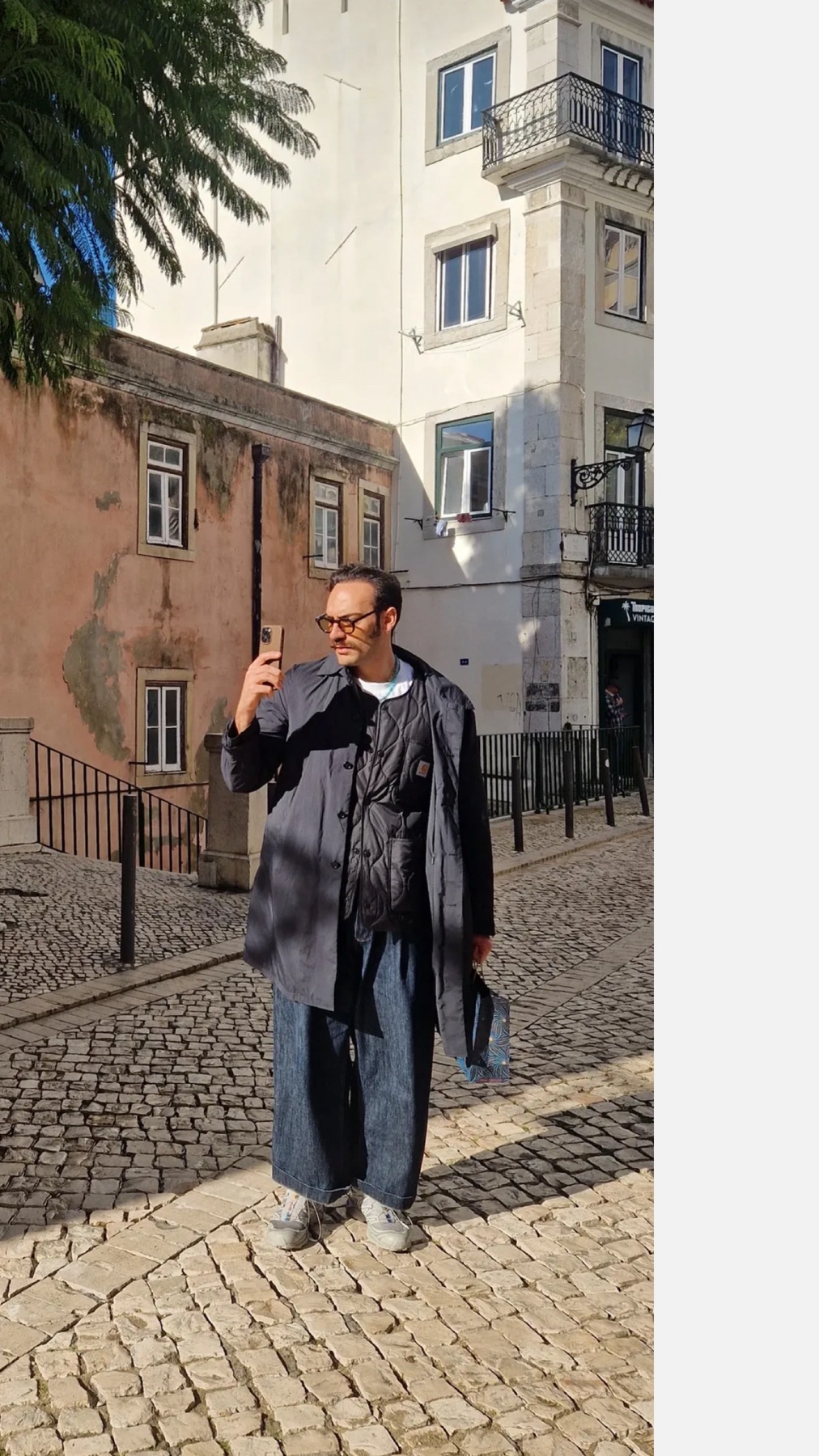 A man poses for a photo in a city street. He is wearing a stylish outfit with glasses and a beard.