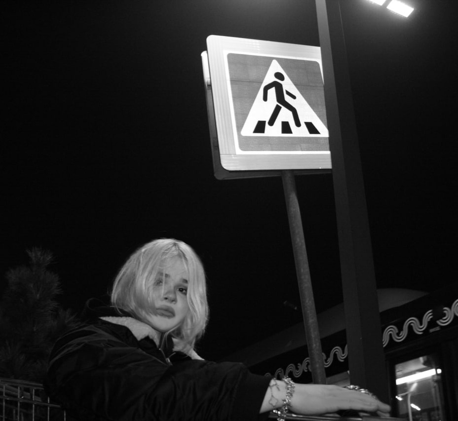 A black and white, low-angle shot of a young woman with blond hair posing at night next to a pedestrian crossing sign. She is wearing a dark jacket and jewelry on her wrist.