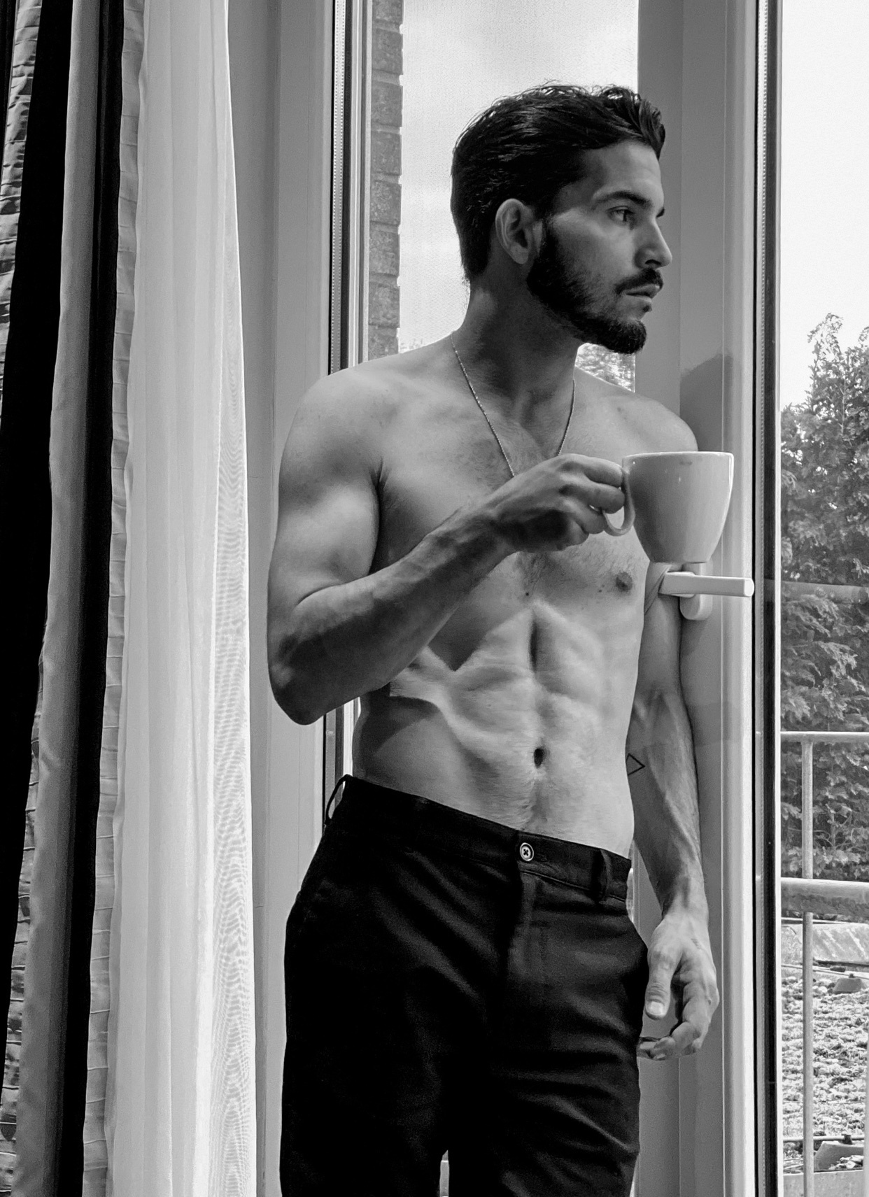 A shirtless man stands by a window, holding a cup of coffee. The black and white photo highlights his physique.