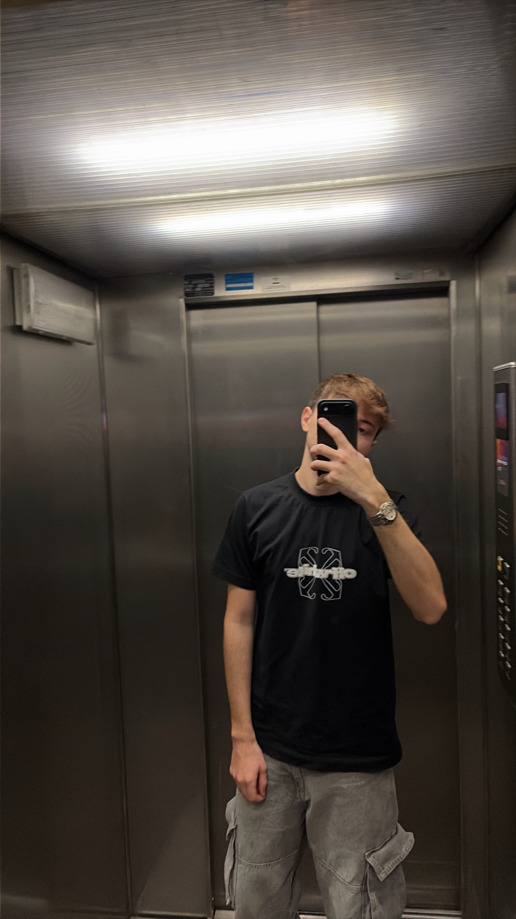 A man takes a mirror selfie in an elevator. He is wearing a black t-shirt and grey cargo pants.