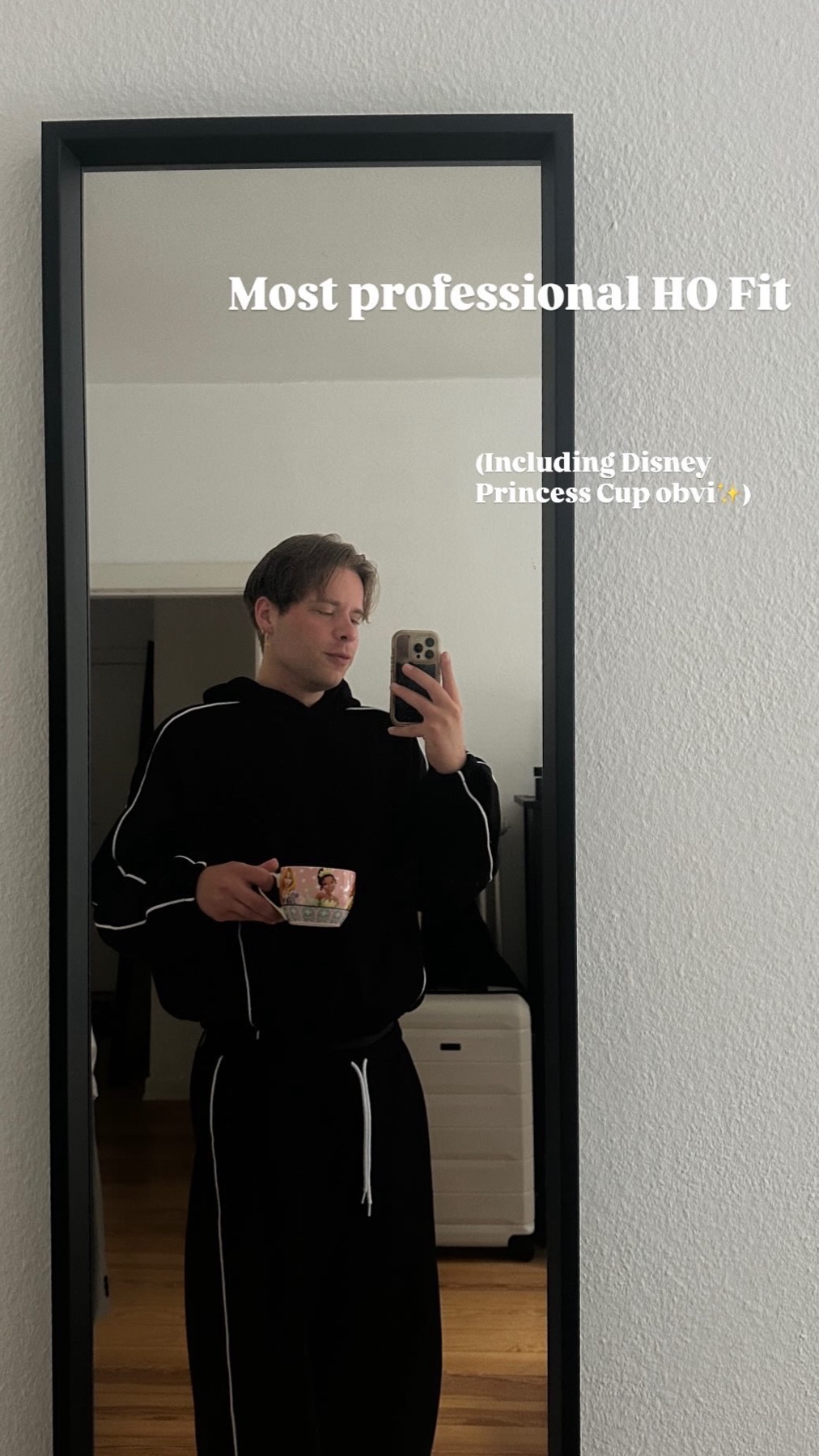 A young man takes a mirror selfie while holding a mug. He is wearing a black hoodie and sweatpants with white trim.
