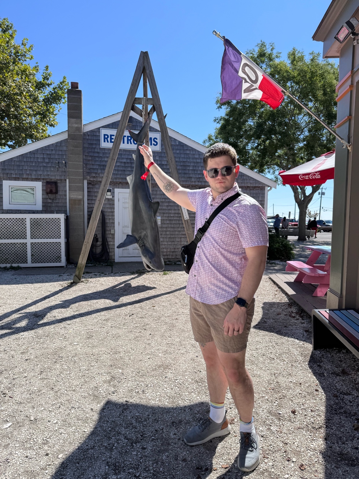 A man in a pink patterned short-sleeved shirt and khaki shorts holds up a large fish he has caught. He is standing in a gravel lot with a building labeled "RESTROOMS" behind him and a flag flying above.