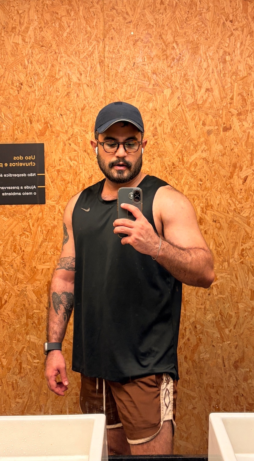 A man with a beard and glasses takes a selfie in a gym locker room. He is wearing a black tank top, brown shorts, and a baseball cap.