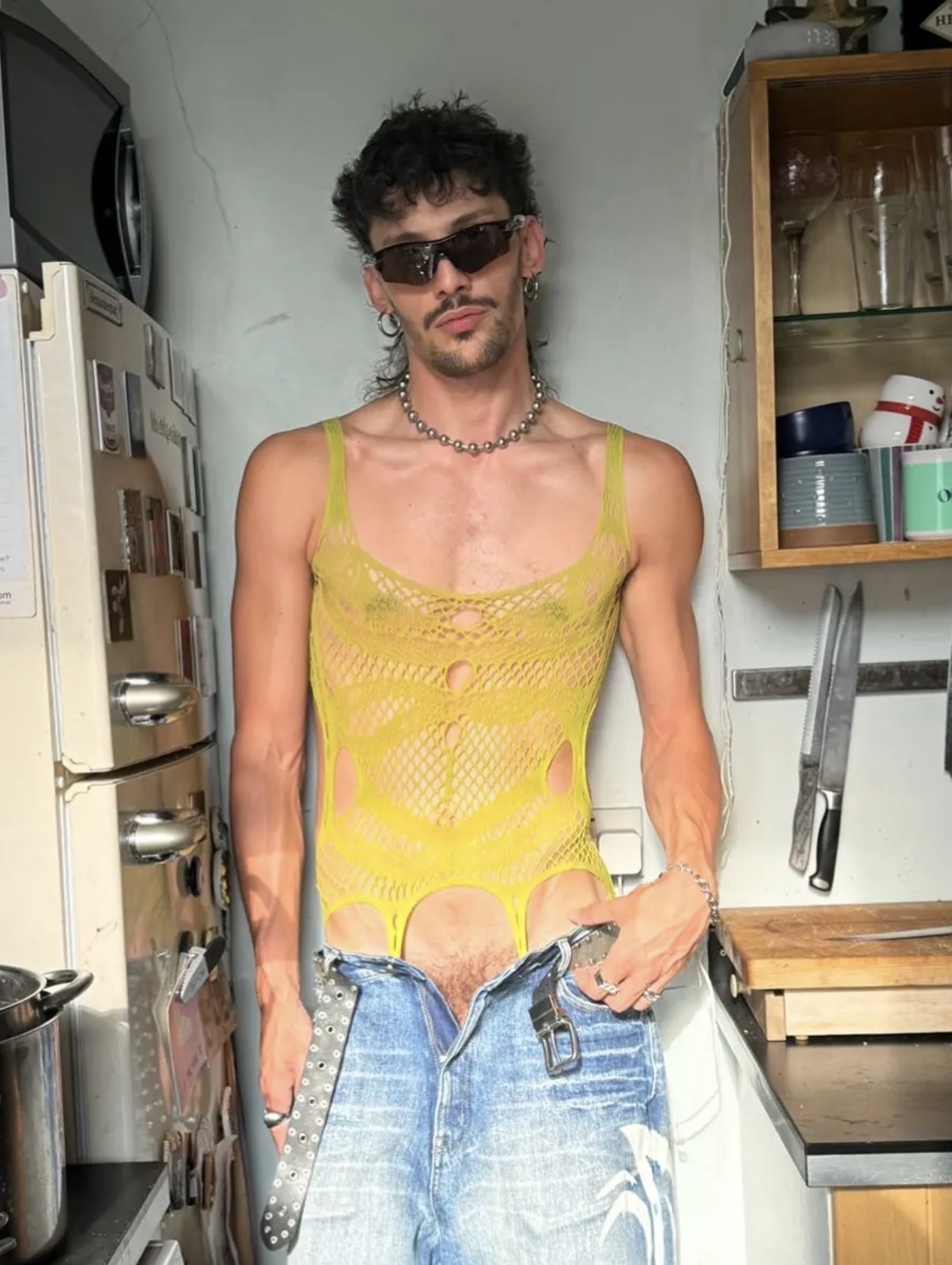 A man with dark, curly hair and sunglasses poses in a kitchen wearing a yellow mesh tank top and low-rise jeans. He is accessorized with a chain necklace and a studded belt.